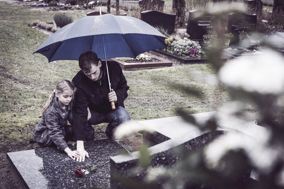 Father and daughter at graveyard visiting grave of mother on rainy day with umbrella